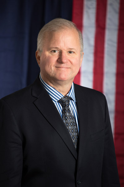 Fotografía de plano medio de Andrew Basiago sonriendo con un traje azul marino, camisa a rayas y corbata estampada.